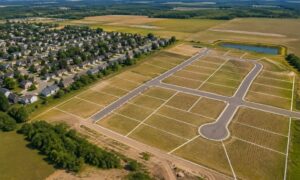 Aerial view of a new housing subdivision with empty rectangular lots and grid streets surrounding a central cul-de-sac, nearby completed homes visible nearby.